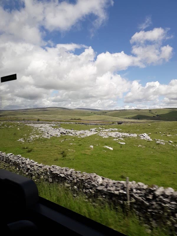 Rolling green hills under a blue sky with fluffy clouds, featuring dry stone walls and scattered rocks in the landscape.