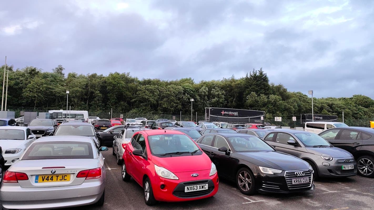 Busy car park with various vehicles, including a red Ford and an Audi, surrounded by greenery and cloudy skies.