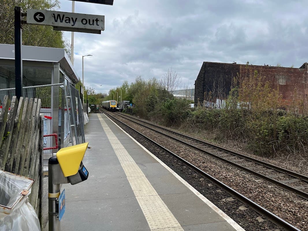 Platform with a "Way out" sign, rail tracks, greenery, and a train approaching in the distance under a cloudy sky.