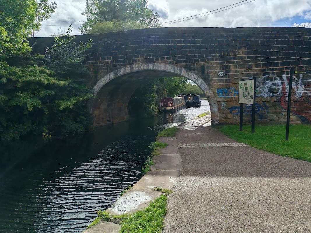 Stone bridge over a canal with moored boats, surrounded by greenery and a pathway beside the water.