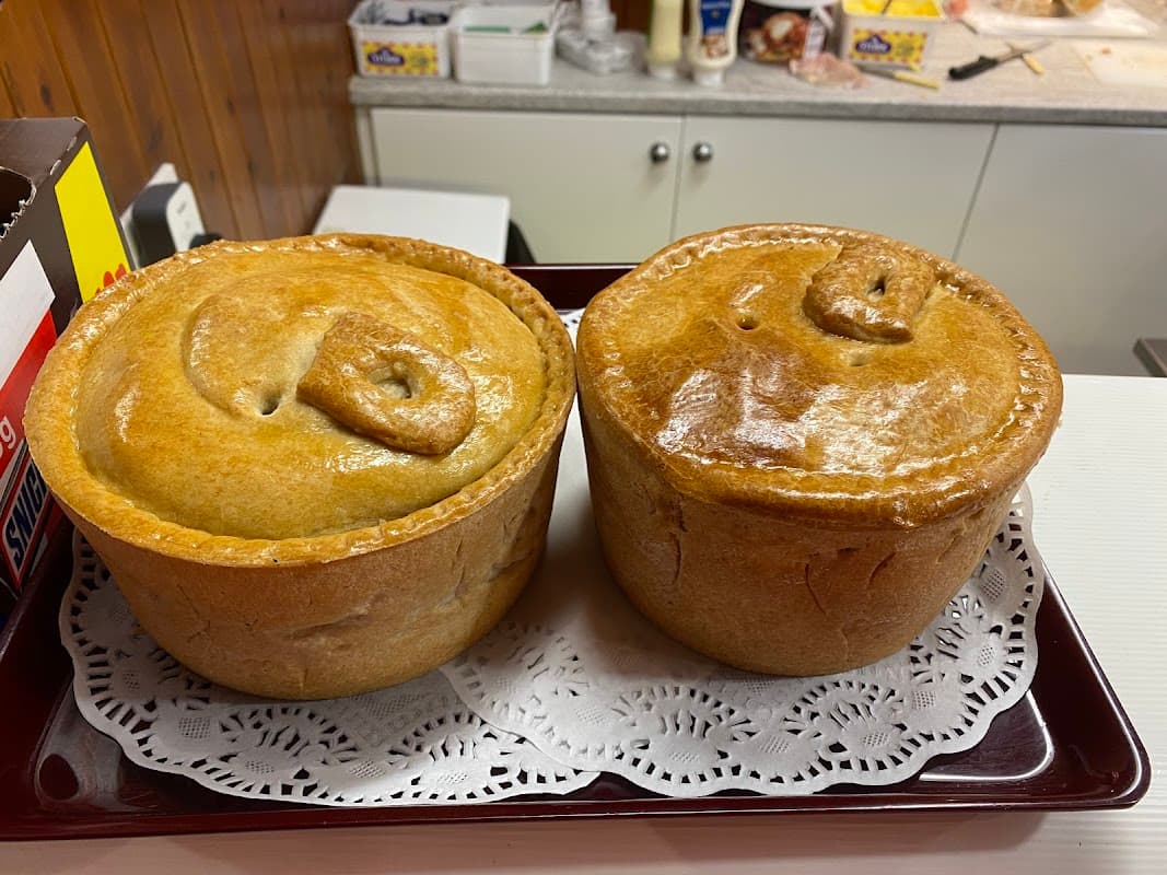 Two golden-brown meat pies on a lace doily, displayed in a bakery setting with various items in the background.