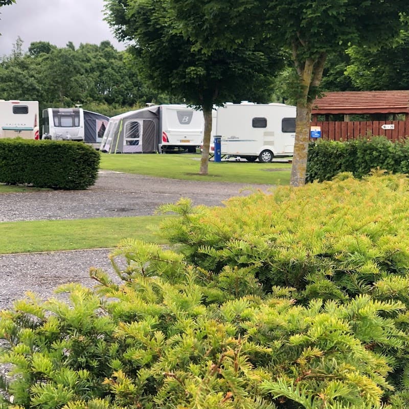 Lush green landscape with neatly trimmed bushes, caravans parked in the background, and trees lining the area.