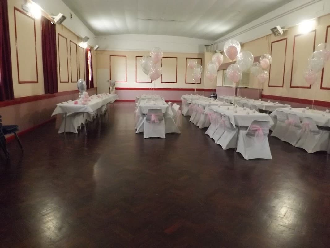 Event venue with tables dressed in white tablecloths and pink bows, decorated with balloons and a polished wooden floor.