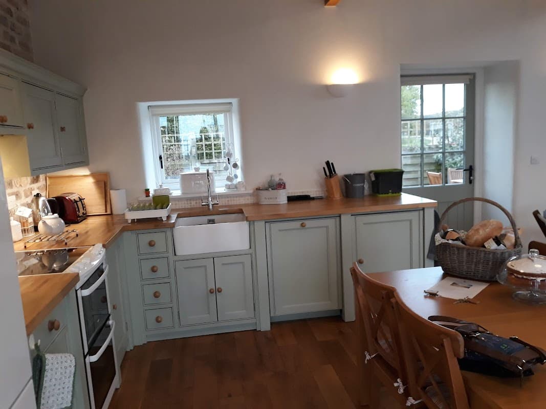 Modern kitchen with light green cabinetry, wooden countertops, and a dining table set with bread and a glass cake stand.