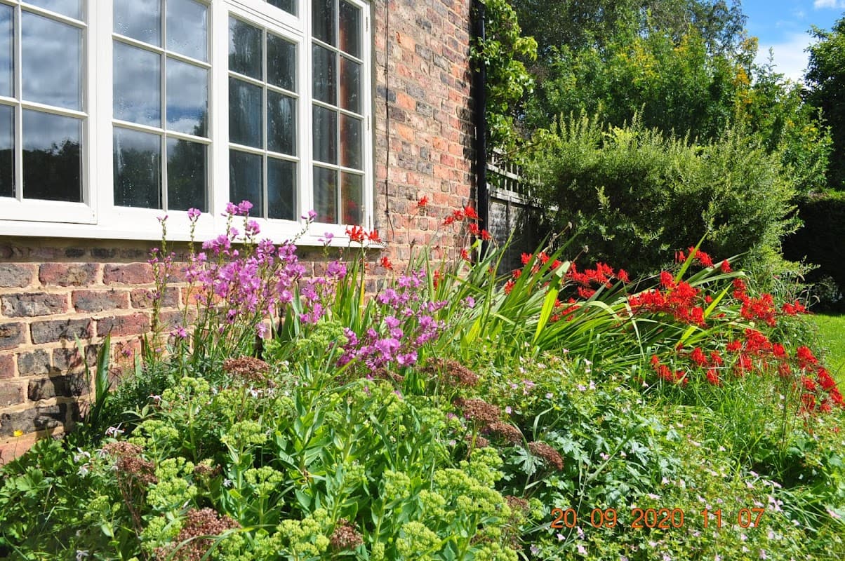 Colorful flower beds with pink and red blooms beside a brick wall and a large window at Warren House Farm Cottages.