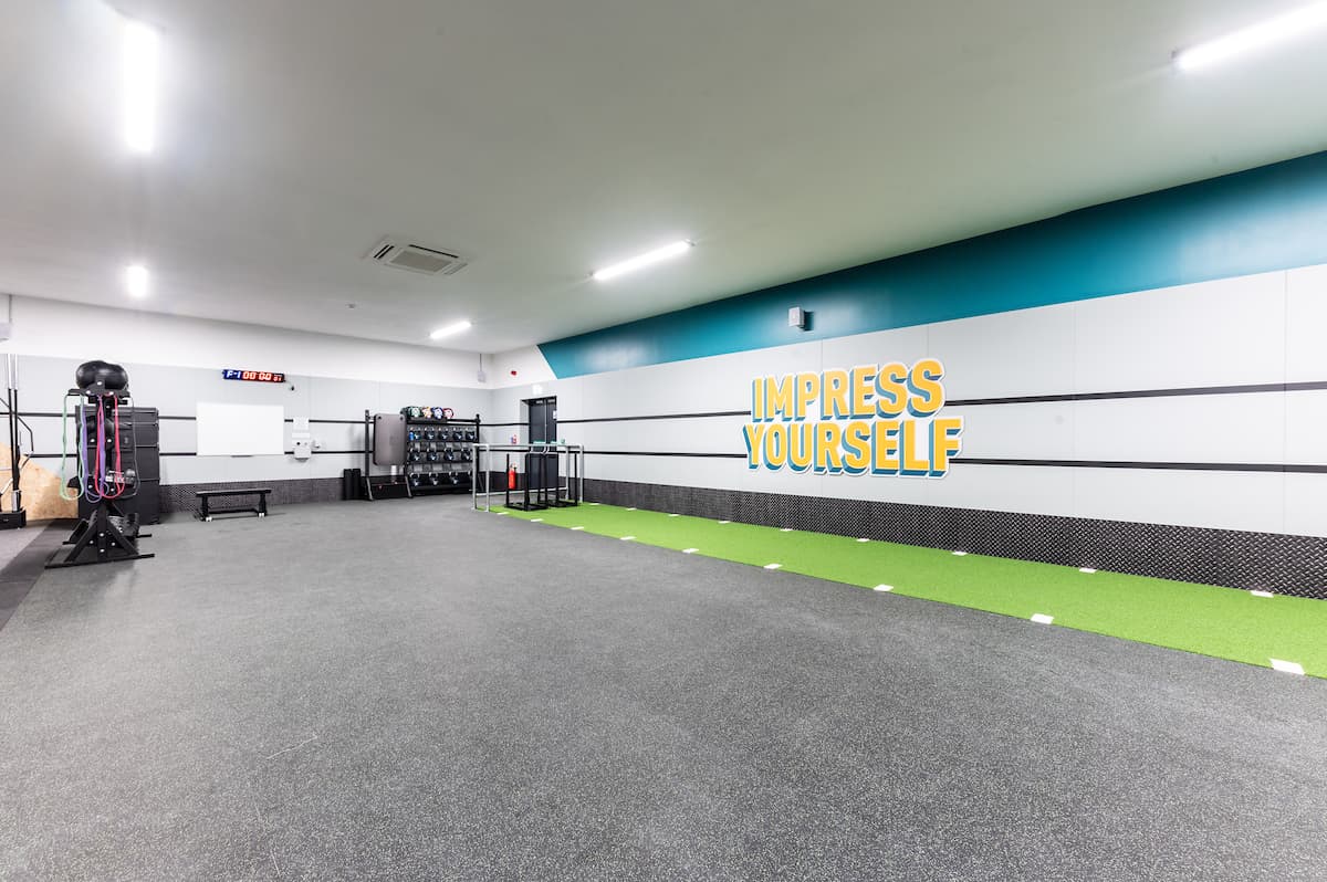 Spacious gym interior with green turf, workout equipment, and a motivational "IMPRESS YOURSELF" wall slogan.