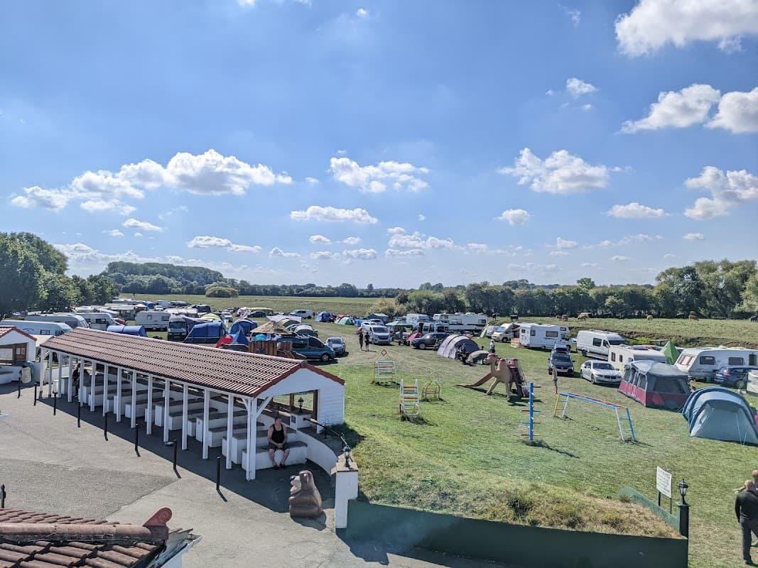 Busy campsite with tents and caravans under a bright blue sky, surrounded by green fields and trees.