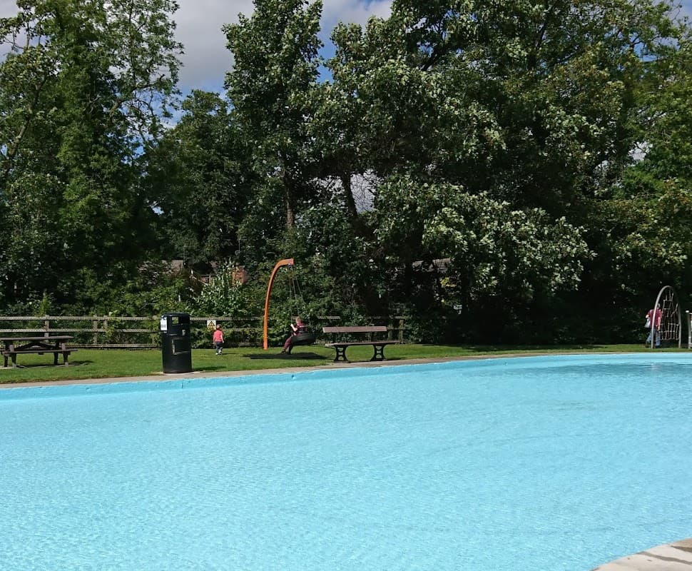 A clear paddling pool surrounded by green trees, benches, and a play area with a slide in Bridge Hewick, Yorkshire.