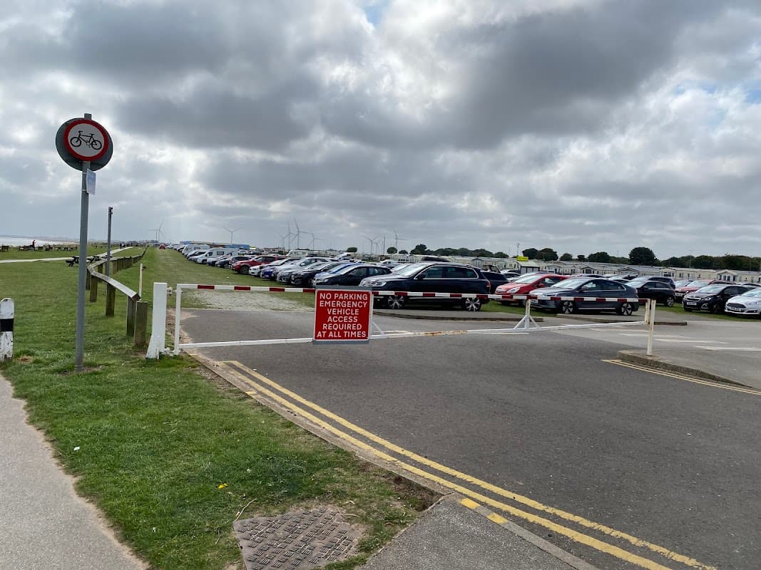 Park and Ride area with parked cars, a no parking sign, and wind turbines in the background under a cloudy sky.