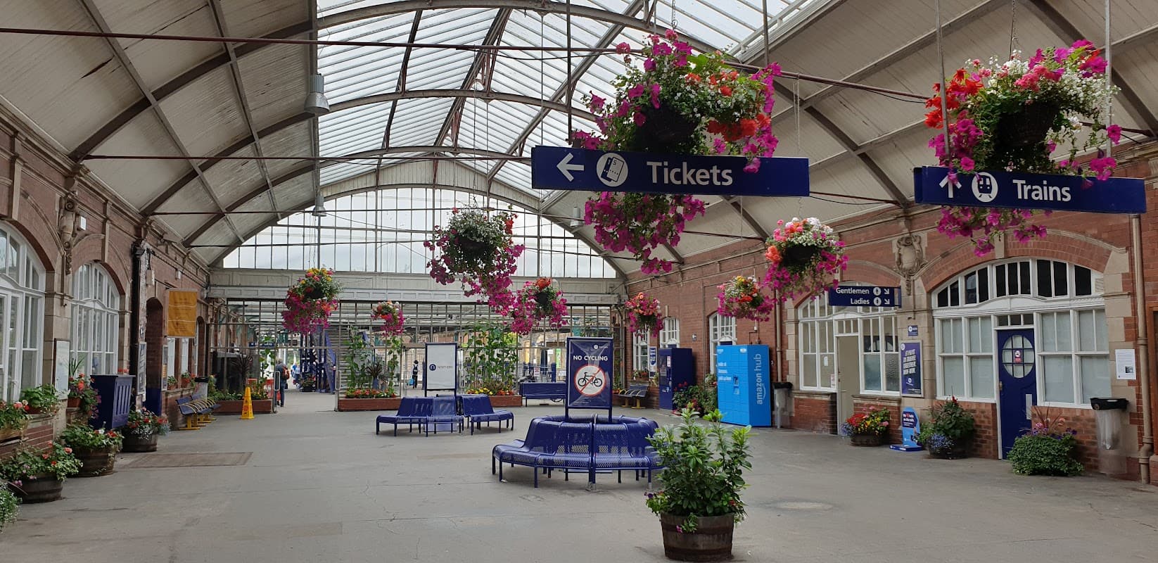 Brightly lit station interior with hanging flower baskets, ticket and train signs, and seating areas.