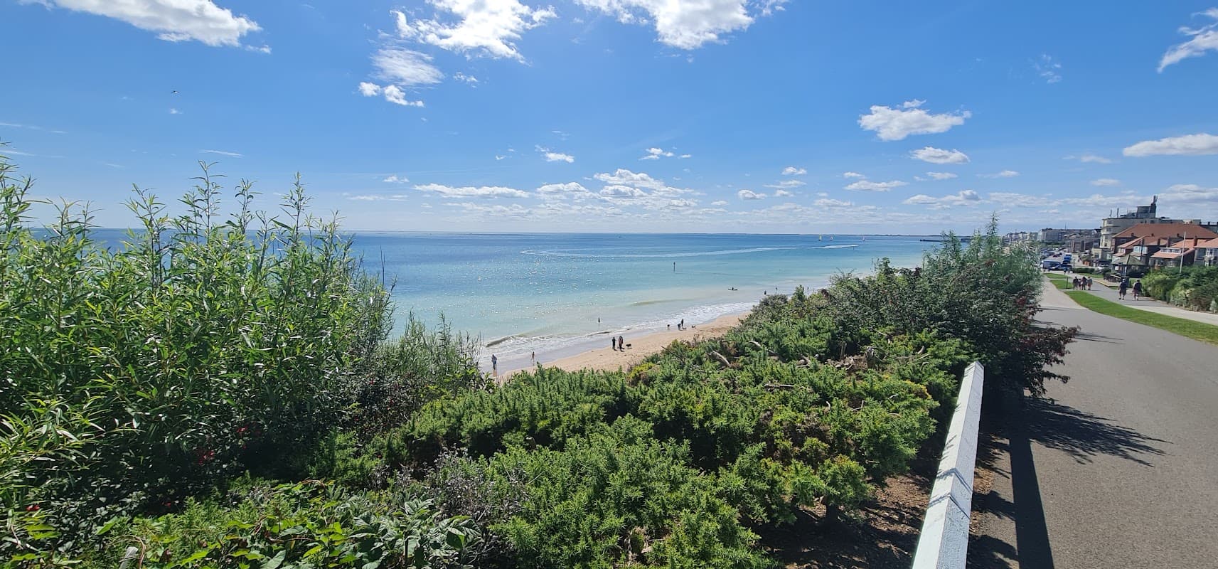 Scenic view of a beach with turquoise water, sandy shore, and people walking, framed by greenery and a clear blue sky.