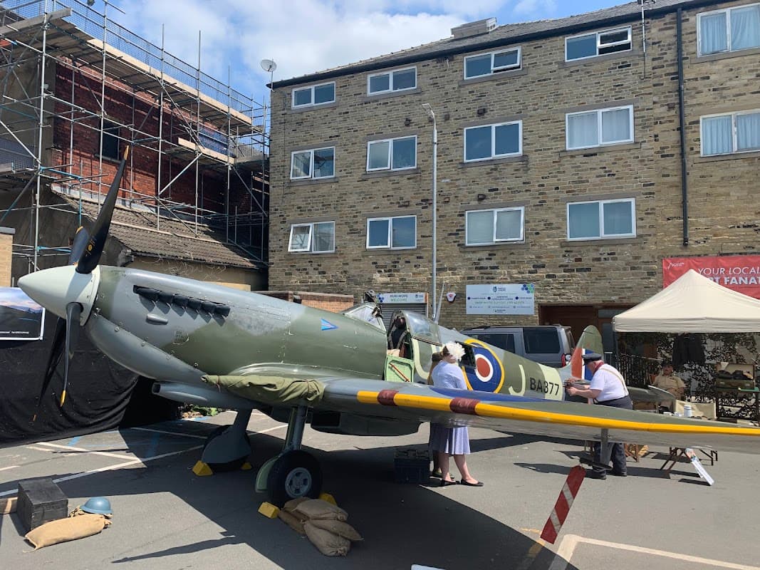 A vintage aircraft displayed in a car park, with nearby scaffolding, buildings, and people interacting around it.