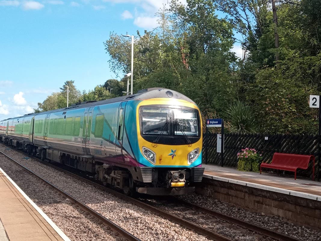 A colorful train at Brighouse station, with greenery and a red bench visible on the platform.