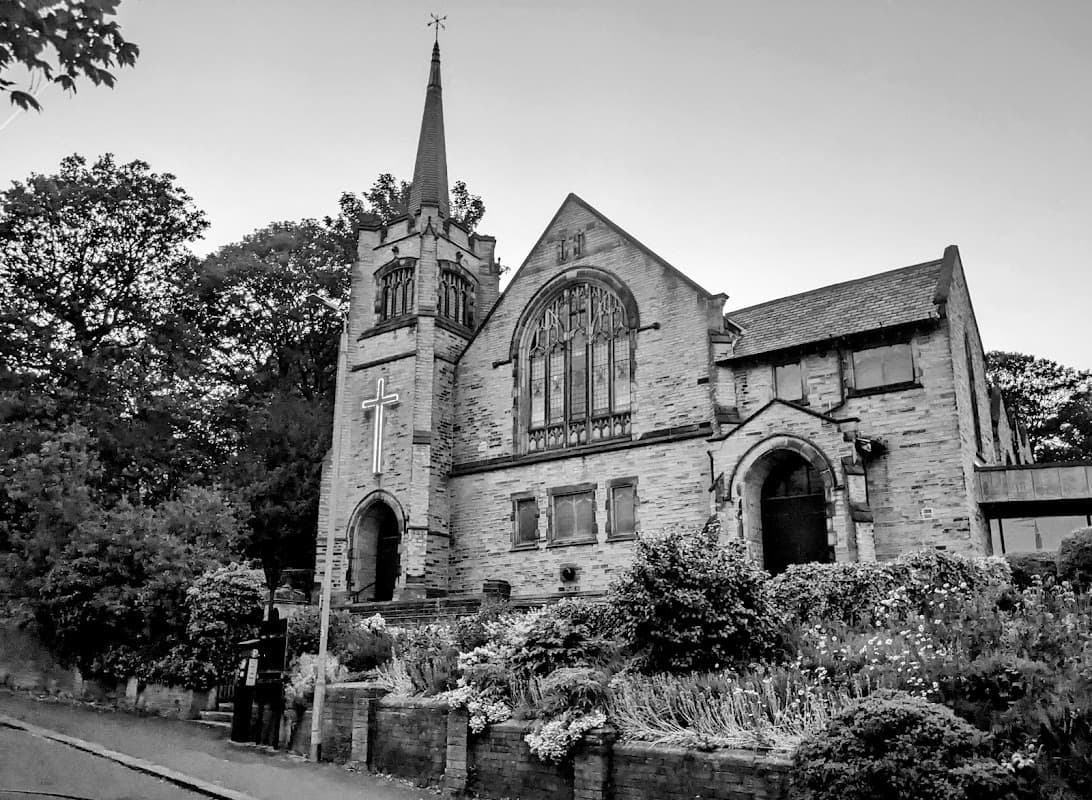 Historic stone church with a tall spire, surrounded by trees and flowering plants, in a quiet setting.