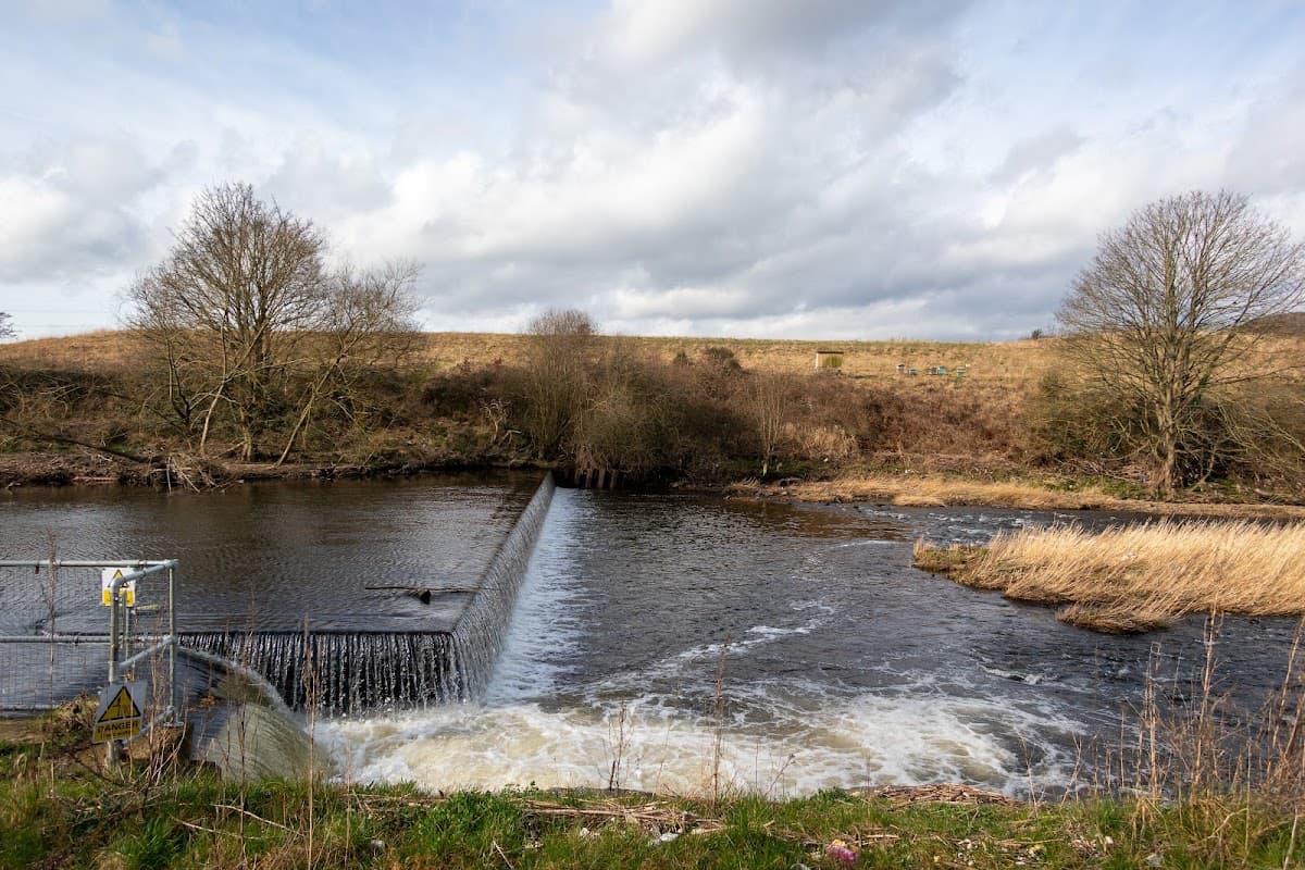 A river with a weir, grassy banks, and trees under a cloudy sky at Cromwell Bottom car park in Brighouse, Yorkshire.
