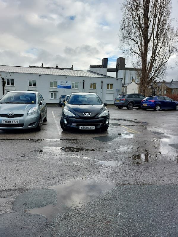 Two parked cars in a gravel lot with puddles, surrounded by buildings under a cloudy sky in Brighouse, Yorkshire.
