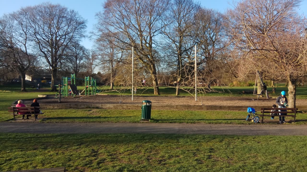 Wellholme Park features playground equipment, benches, trees, and a grassy area under a clear blue sky.