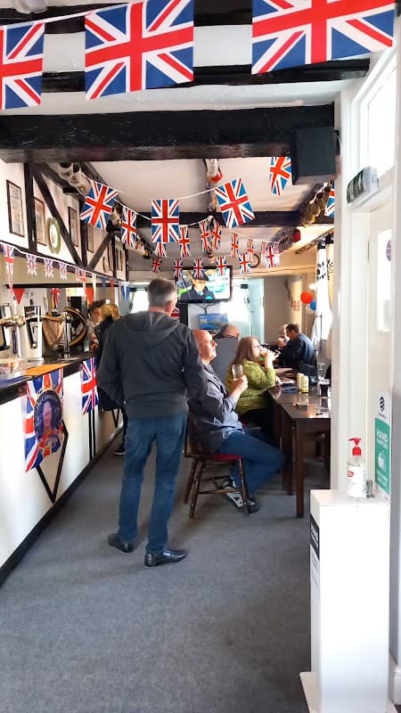 A cozy pub interior decorated with Union Jack flags, featuring patrons enjoying drinks and a TV in the background.