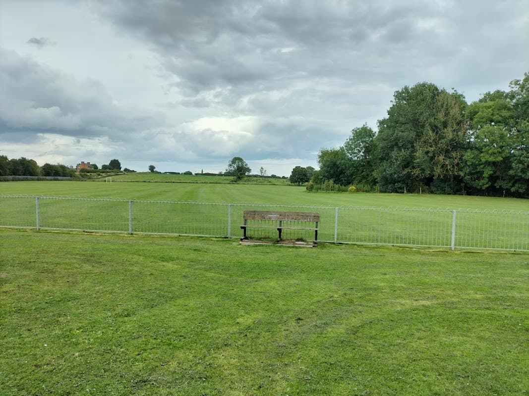 A grassy field with a wooden bench in front of a fence, under a cloudy sky in Brompton, Yorkshire.