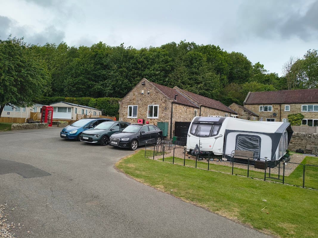 Caravan park with parked cars, a caravan, stone buildings, and lush greenery under a cloudy sky.