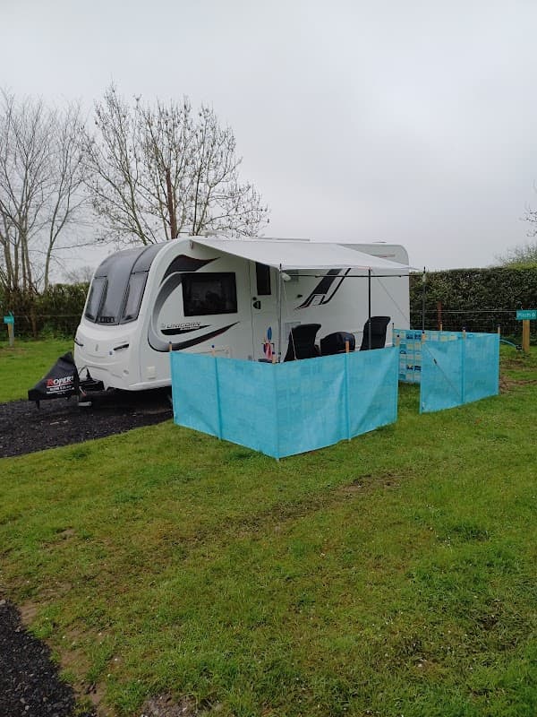 A caravan with an awning set up on a grassy site, surrounded by blue windbreaks and trees in the background.