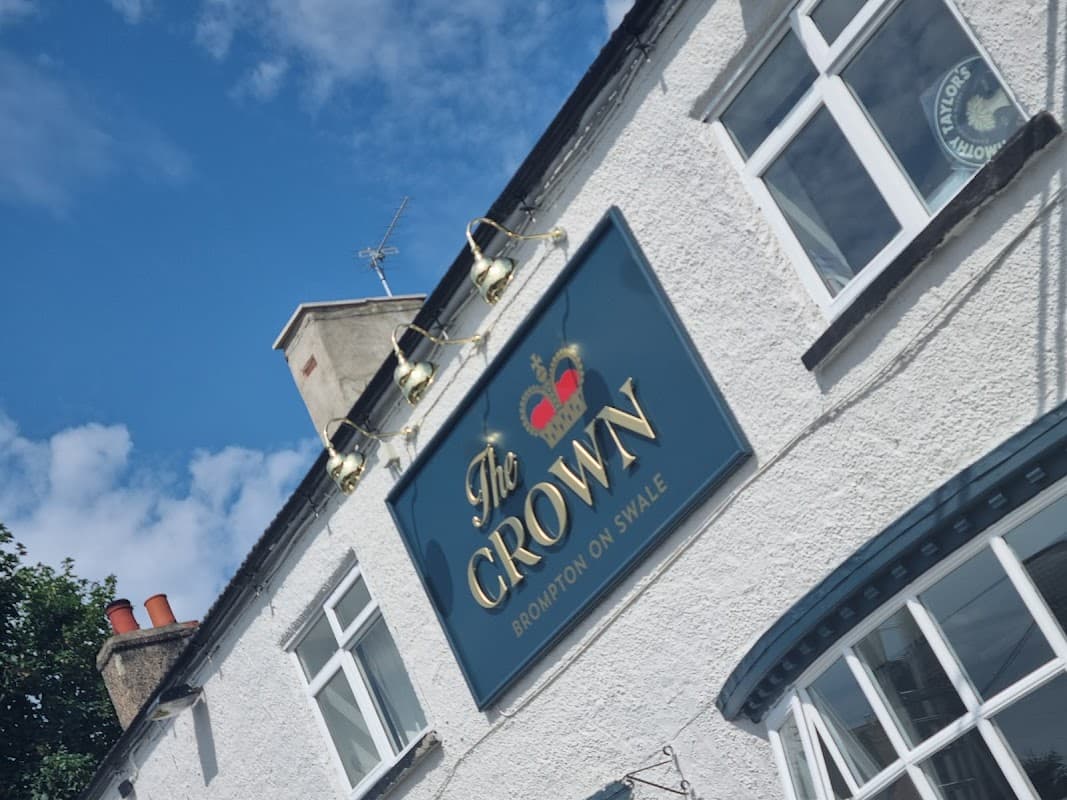 The Crown pub sign with gold lettering, blue background, and a crown logo, set against a clear blue sky.