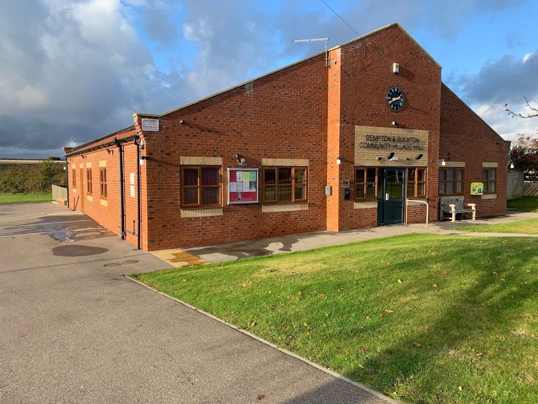 Brick building with a clock, large windows, and community notice board, set against a cloudy sky. Green lawn in front.