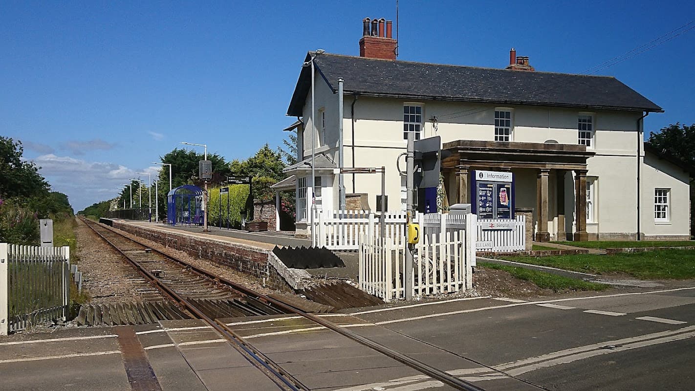 Victorian-style train station building with a platform, surrounded by greenery and a clear blue sky.