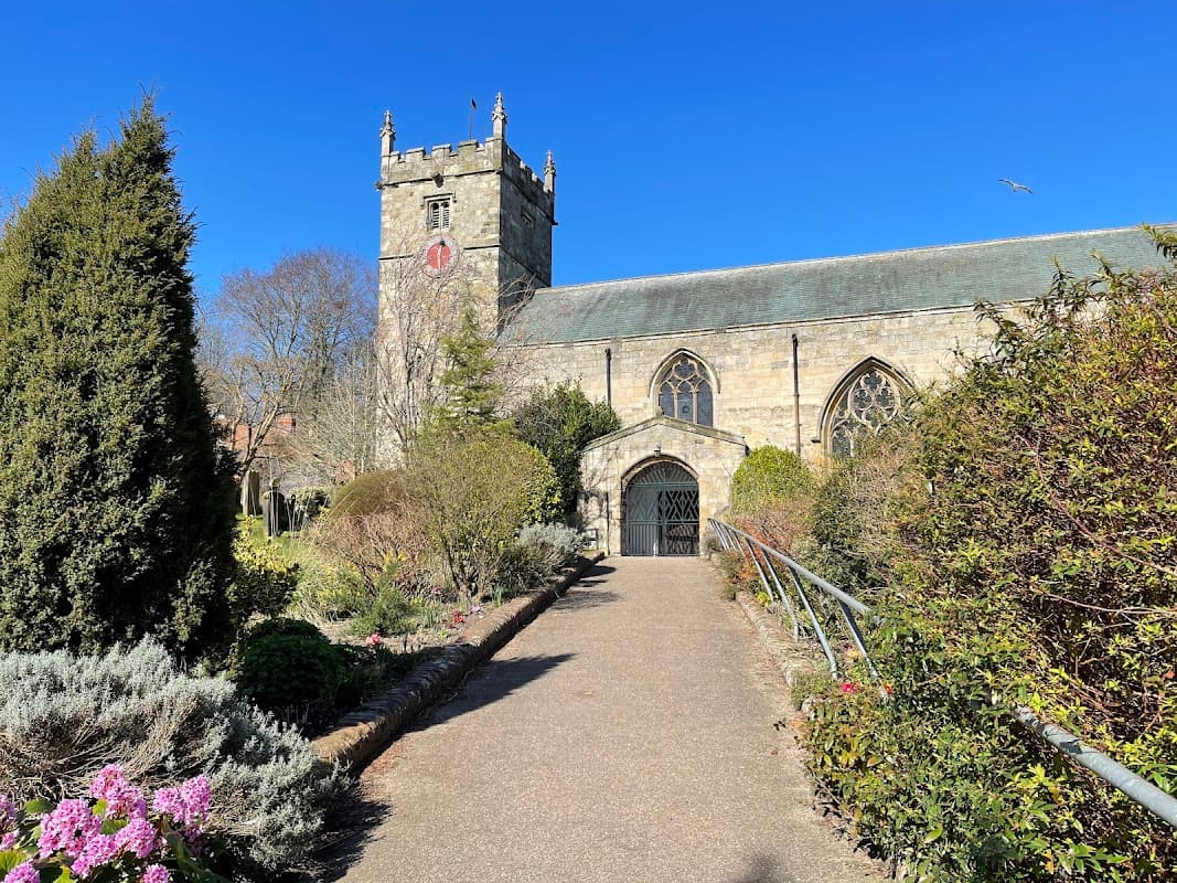 Historic church with a tower, surrounded by lush greenery and colorful flowers under a clear blue sky.