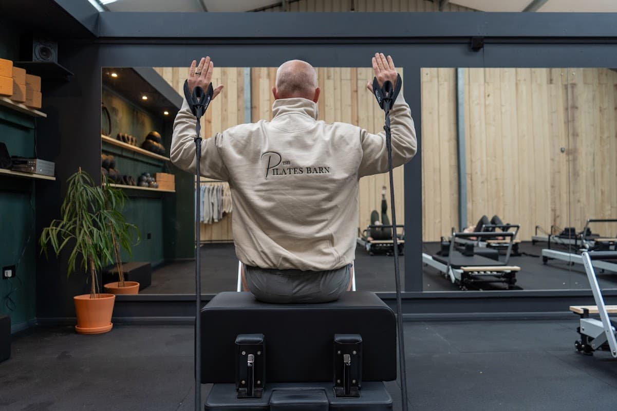 A person in a Pilates jacket sits on a reformer, facing a mirror in a well-lit studio with exercise equipment.