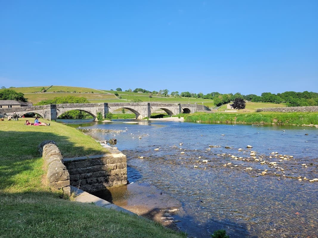 Scenic view of a stone bridge over a river, lush green hills, and a grassy area with people relaxing.