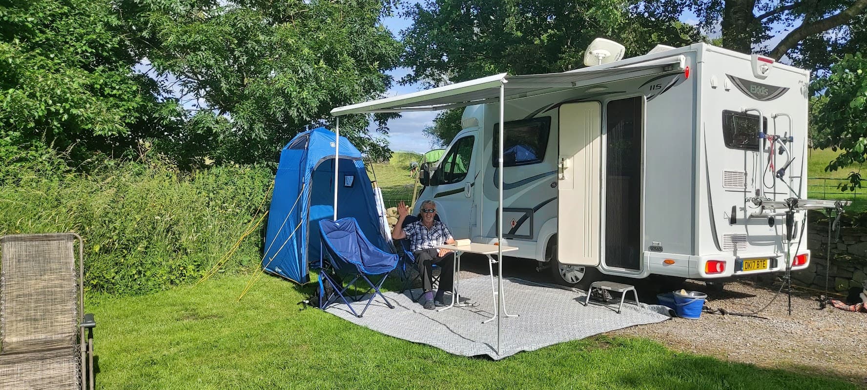 A camper van with a blue tent nearby, set on green grass, with a person sitting at a table under a canopy.