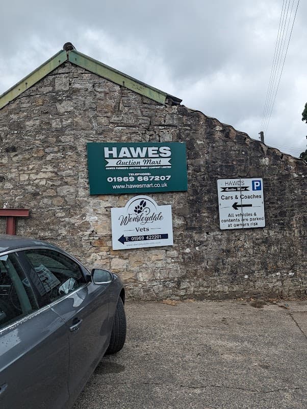 Hawes Farmers' Auction Mart sign on a stone building, with parking and directional signs for cars and coaches.