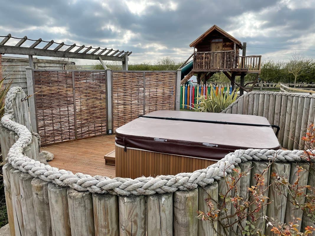 Hot tub enclosed by wooden fencing, with a treehouse and slide in the background under a cloudy sky.