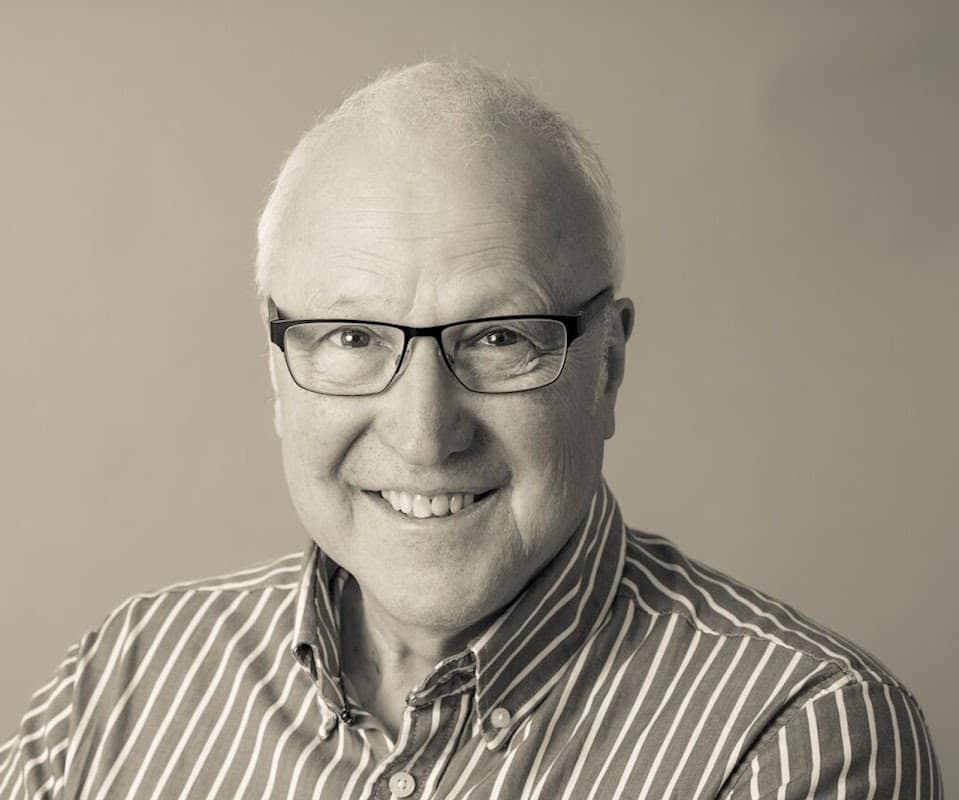 Smiling older man with glasses, wearing a striped shirt, against a neutral background.