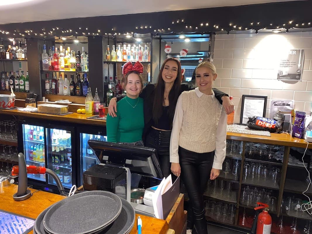 Three women stand behind a bar with shelves of liquor and festive decorations, smiling at the camera.