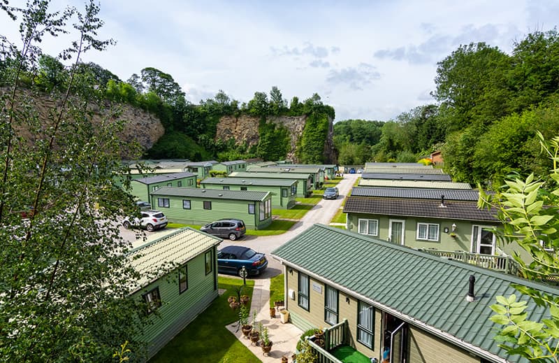 Aerial view of Low Bridge Holiday Park featuring green holiday homes surrounded by trees and a rocky cliff backdrop.