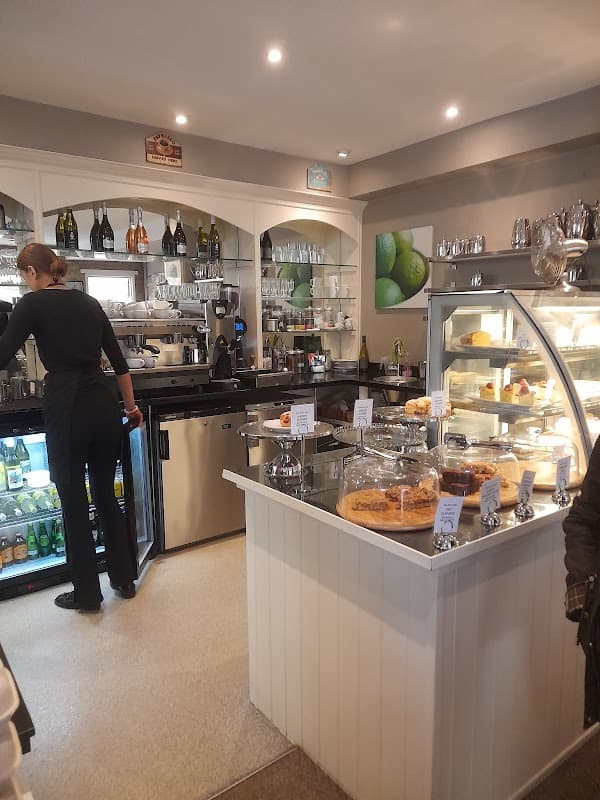 Cozy cafe interior with a counter displaying cakes, drinks, and a barista preparing coffee. Green lime decor visible.