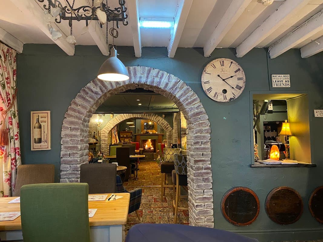 Cozy bar interior with exposed brick arches, wooden beams, a clock, and warm lighting, featuring a fireplace and seating.