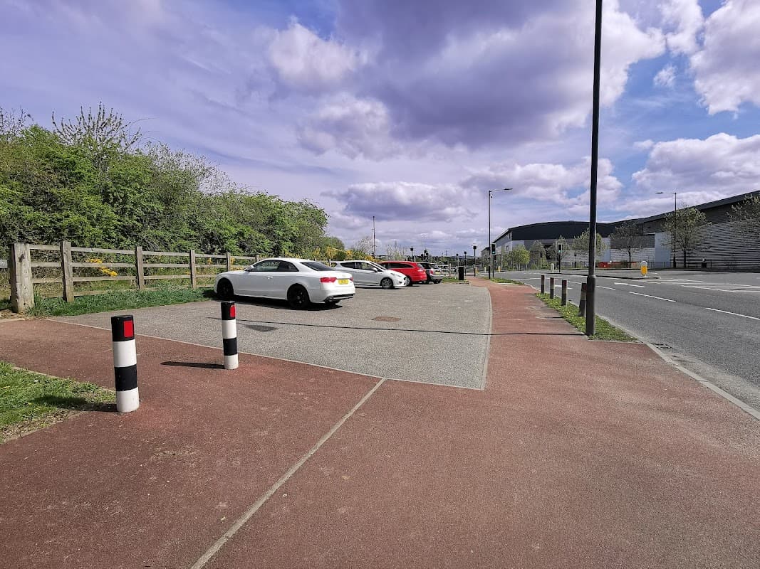 Several parked cars in a gravel car park, surrounded by greenery and a pathway, under a partly cloudy sky.