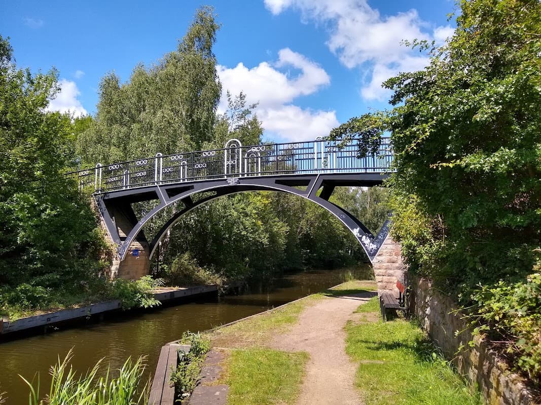 Black iron bridge arching over a calm canal, surrounded by lush greenery and a walking path.