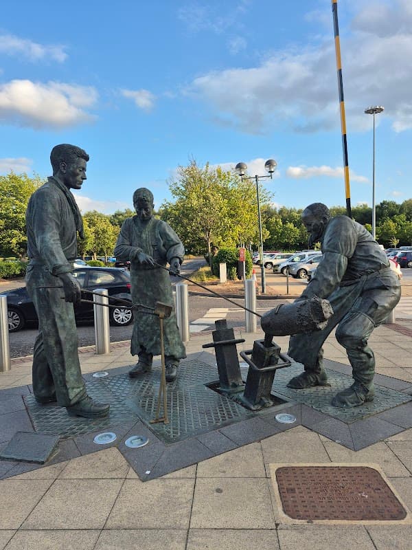Bronze statue of three workers in a car park, surrounded by trees and parked cars under a blue sky.