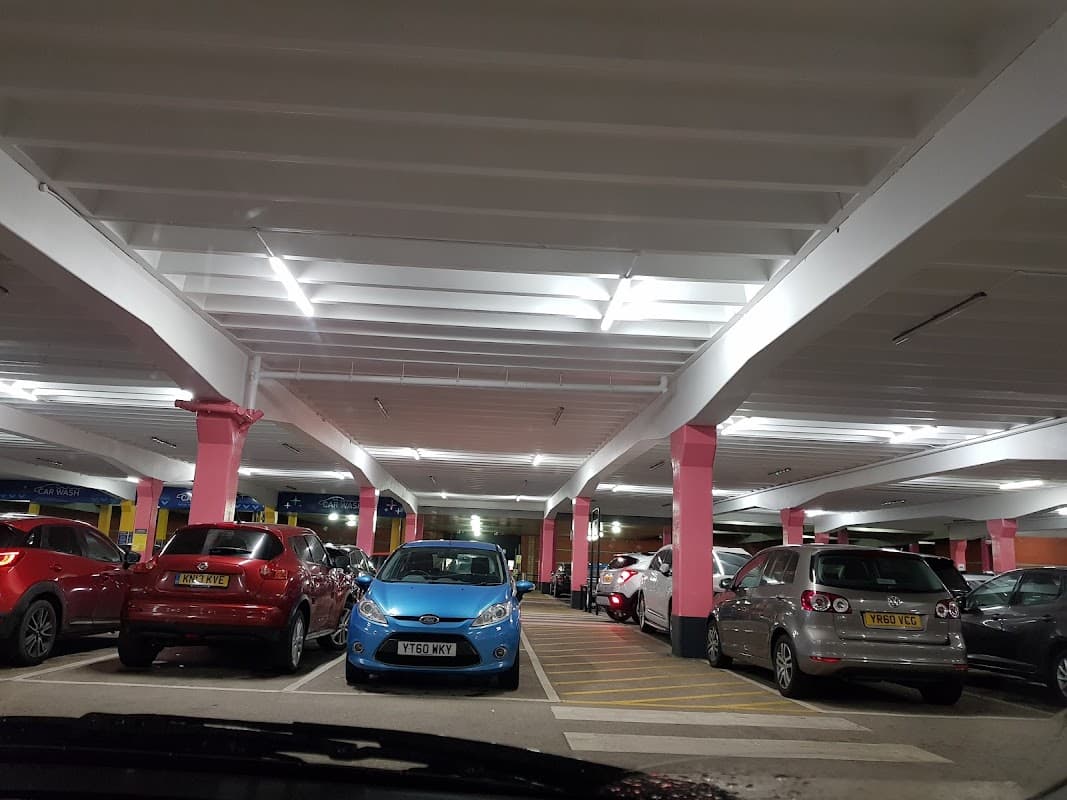 Brightly lit car park with pink columns, featuring various parked cars including a blue one.