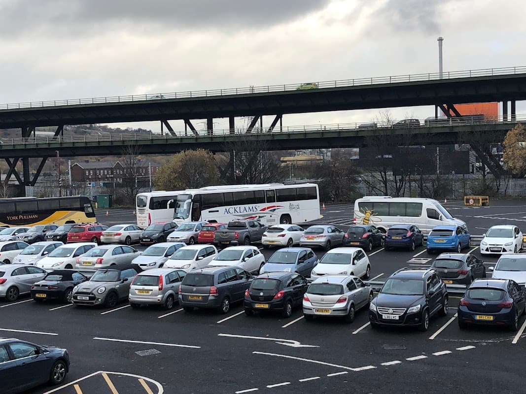 Meadowhall staff car park filled with cars and buses, under a railway bridge, cloudy sky overhead.