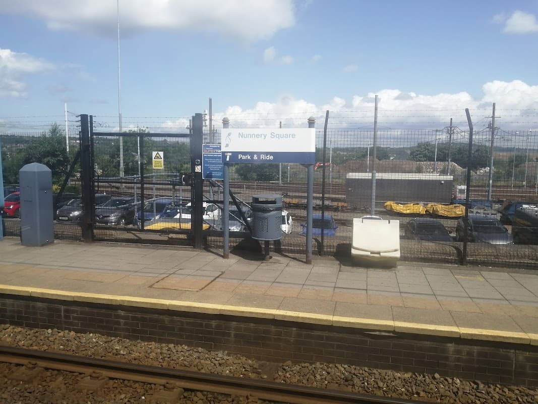 Sign for "Nunnery Square - Park & Ride" with parked cars and a fenced area in the background under a blue sky.
