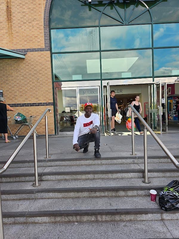 A man in a white sweatshirt kneels on steps outside a shopping center with shoppers entering and exiting.