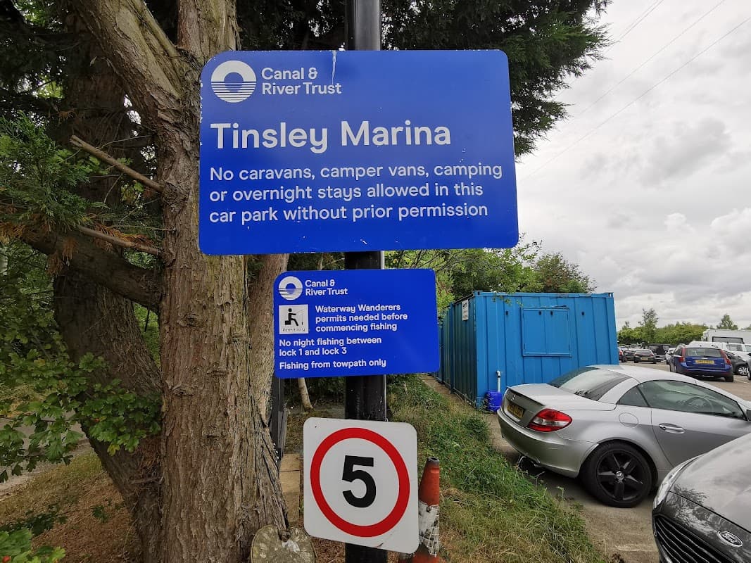 Sign for Tinsley Marina car park with rules, blue container in background, and a parking sign indicating a speed limit.