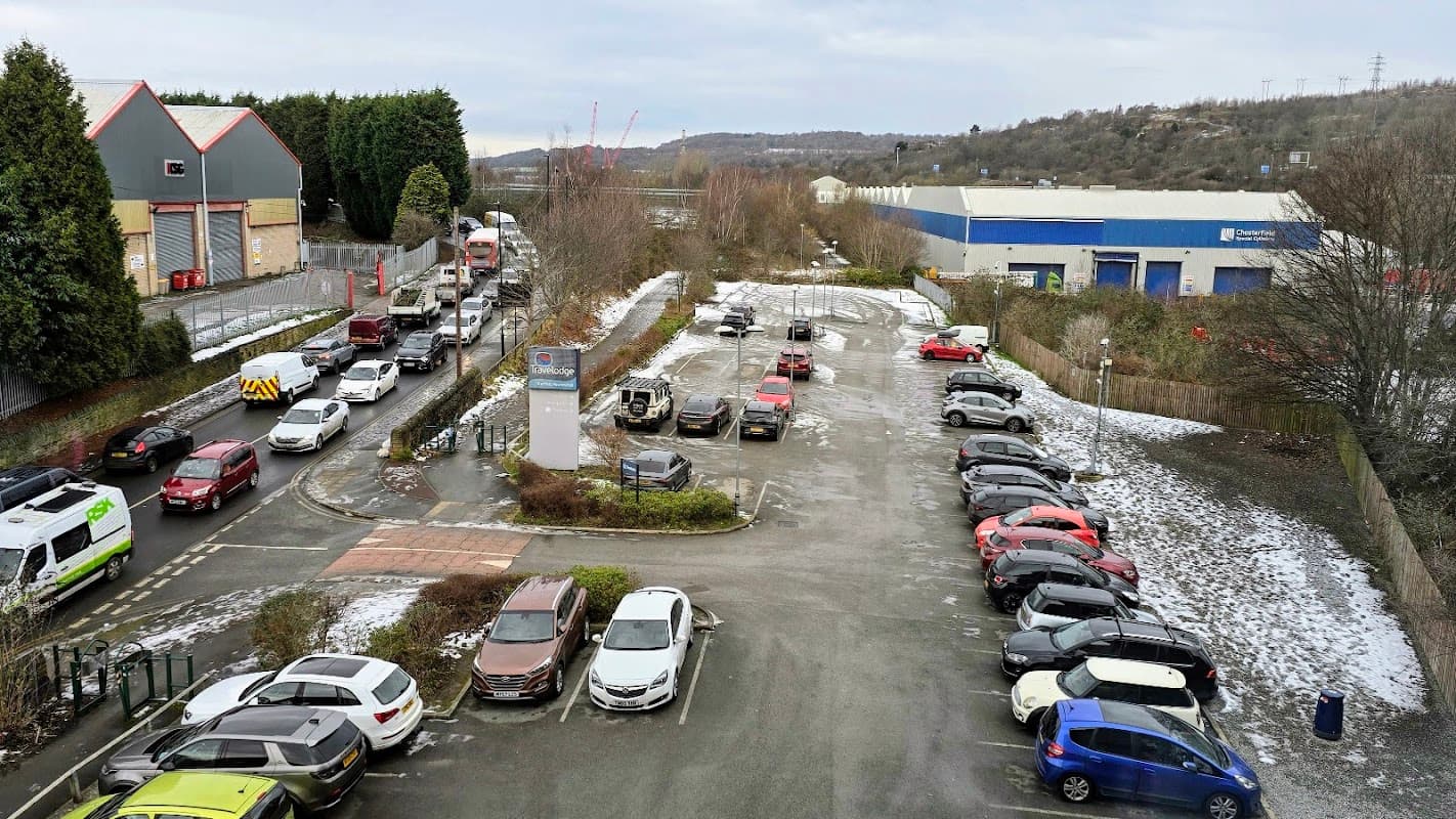 Travelodge car park with parked cars, snow patches, and surrounding greenery; industrial buildings in the background.