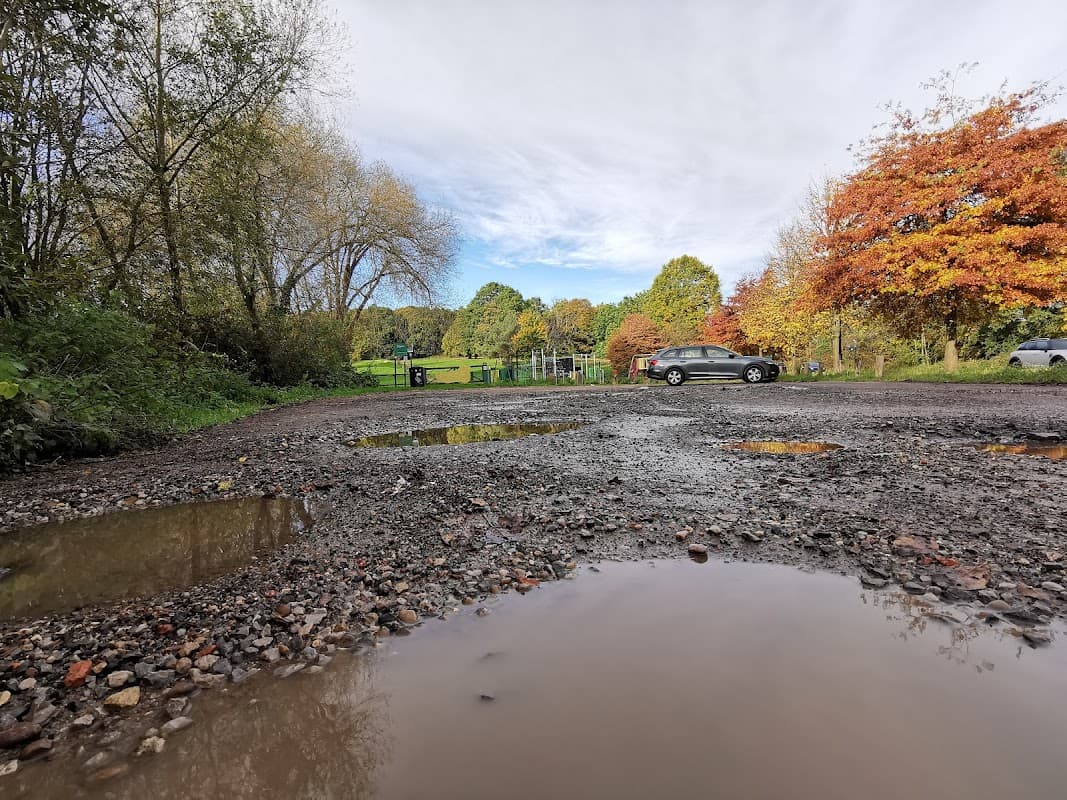 Gravel car park with puddles, surrounded by trees displaying autumn colors and a distant green field.