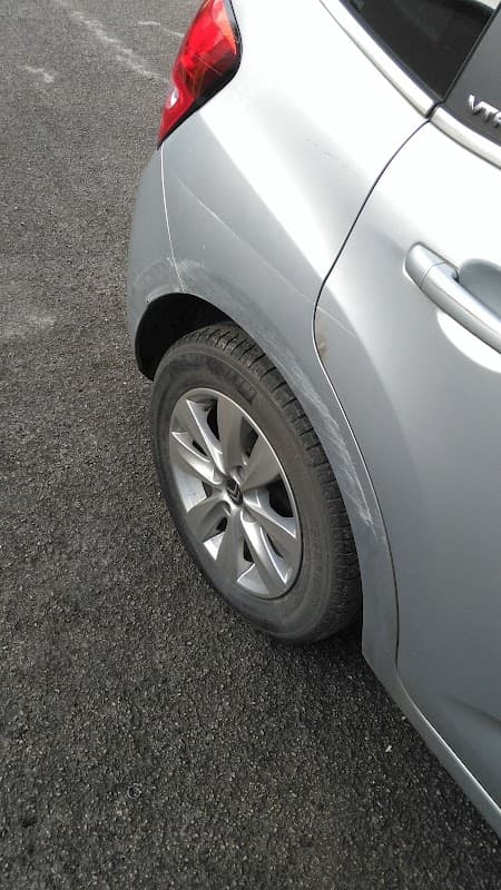 Silver car with a damaged rear wheel and tire on a gravel surface outside a body repair shop.